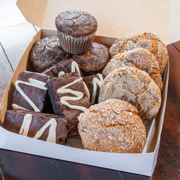 Open pastry box filled with chocolate muffins, sugar-coated cookies, and chocolate brownies drizzled with white icing.