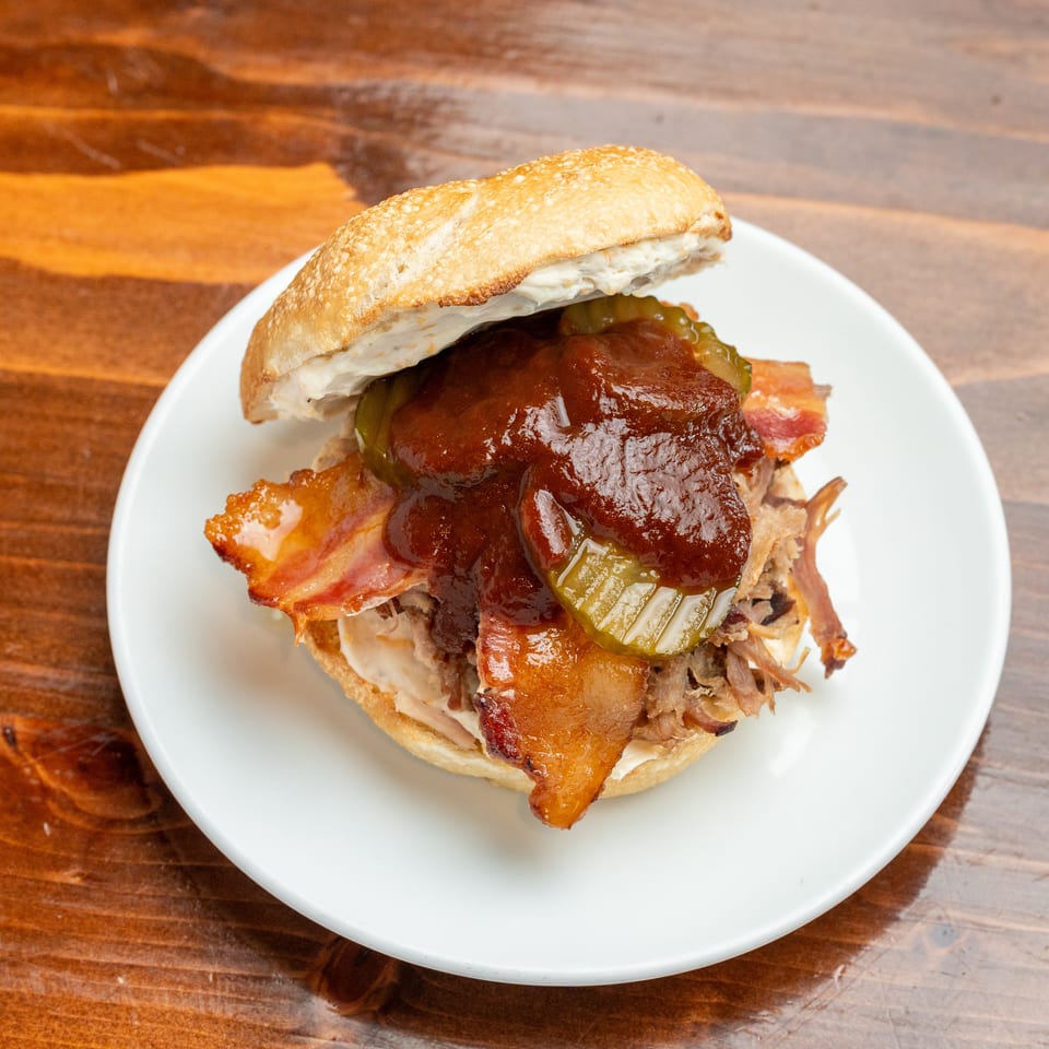 Loaded barbecue sandwich with bacon, pickles, creamy sauce, and shredded meat on a white plate atop a wooden table.