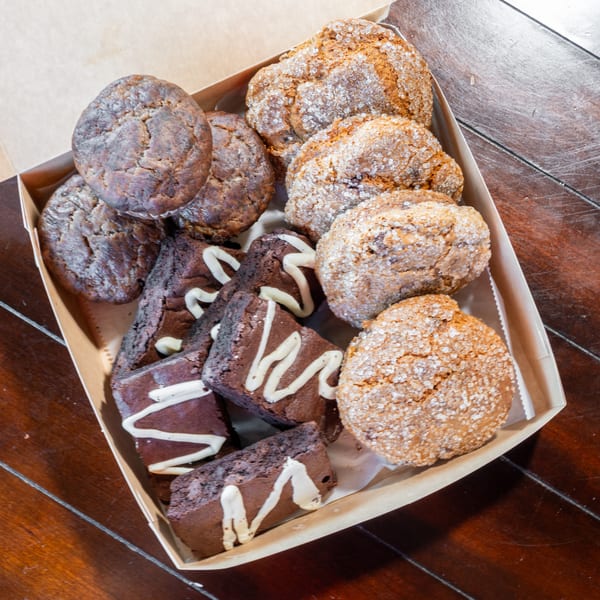 Assorted chocolate brownies, cookies, and sugar-coated pastries arranged in a cardboard bakery box on a wooden table.