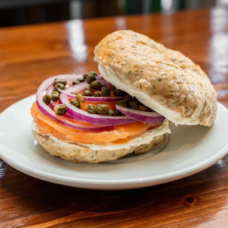 Bagel sandwich layered with smoked salmon, cream cheese, tomato, red onion, and capers on a white plate.