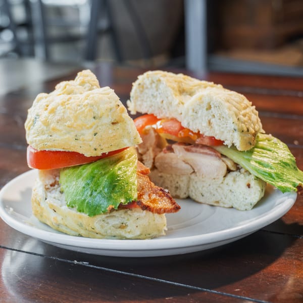 Two toasted chicken and bacon sandwiches with lettuce and tomato on a white plate atop a wooden restaurant table.