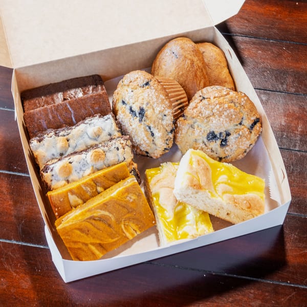 Open bakery box filled with assorted muffins, blueberry pastries, chocolate brownies, loaf cakes, and lemon-glazed slices.