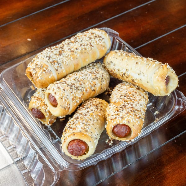A tray of sesame- and seed-topped sausage rolls sits on a wooden table in a clear plastic container.