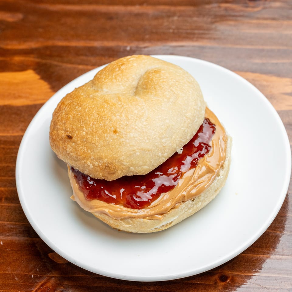 A golden bun sandwich filled with peanut butter and red jam sits on a white plate atop a wooden table.