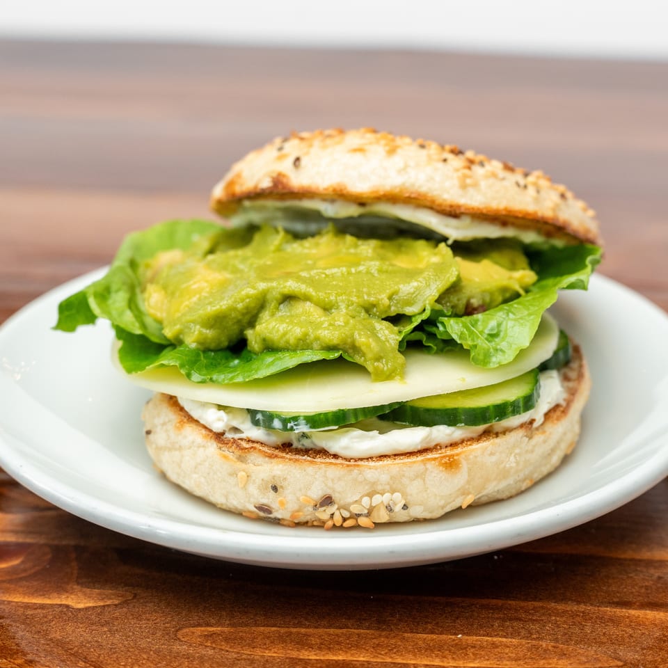 Open-faced sandwich on a seeded bun with lettuce, avocado, cheese, cucumber, and creamy spread, served on a white plate.