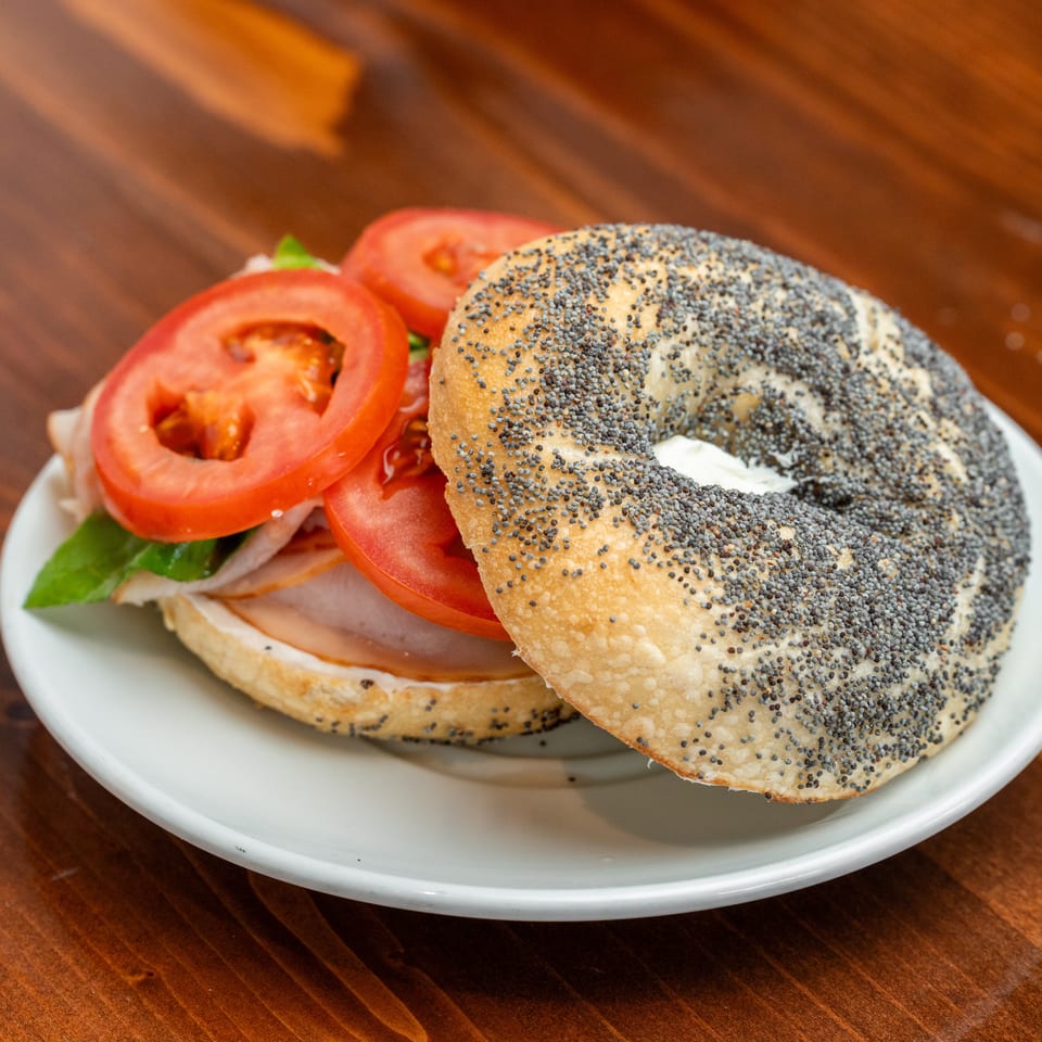 Poppy seed bagel sandwich with sliced tomatoes, lettuce, onions, and deli meat on a white plate.