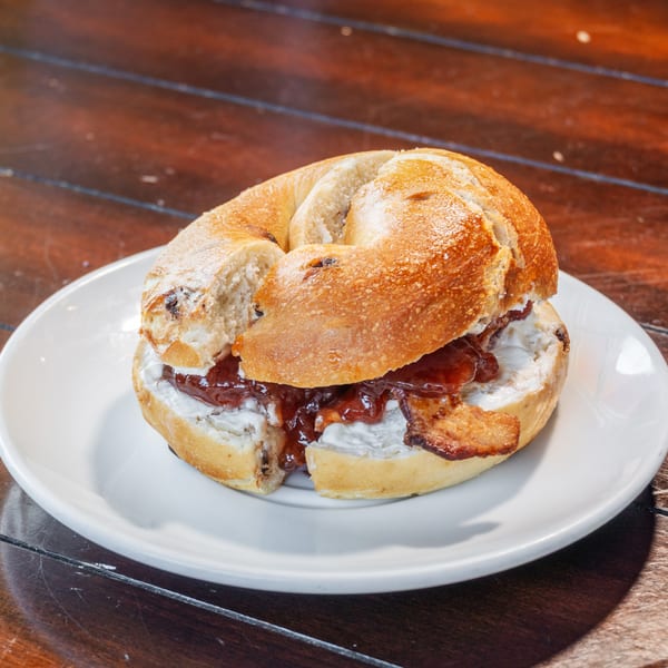 Toasted bagel sandwich with cream cheese, red jam, and bacon served on a white plate atop a wooden table.