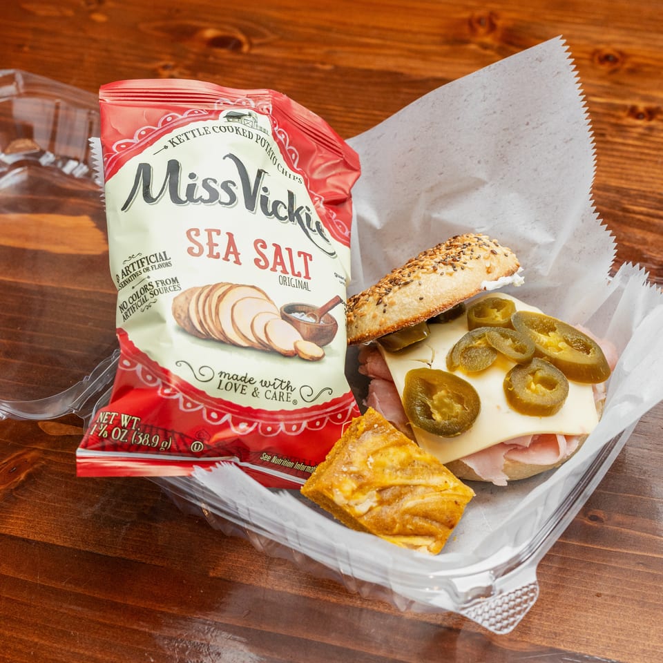 Takeout tray with a jalapeño and ham sandwich, triangular toasted snack, and Miss Vickie’s sea salt potato chips on a wooden table.