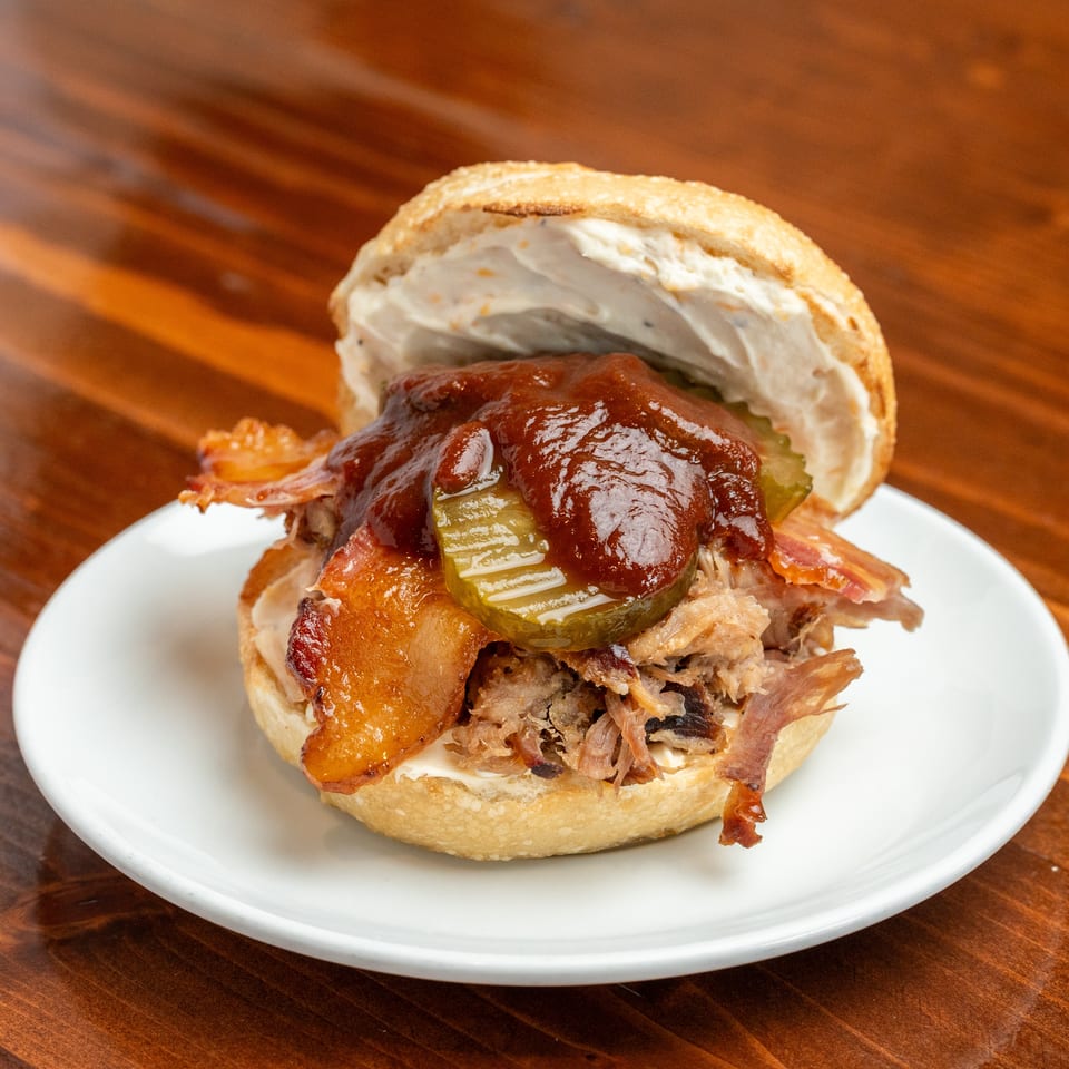 Pulled pork sandwich with bacon, pickles, barbecue sauce, and creamy dressing on a bun, served on a white plate.