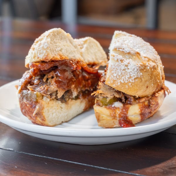 Three powdered-sugar-dusted sandwiches filled with saucy shredded meat sit on a white plate atop a wooden table.