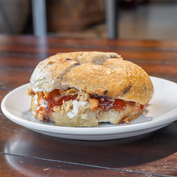 Toasted bagel sandwich layered with cream cheese, red jam, and crunchy topping on a white plate.