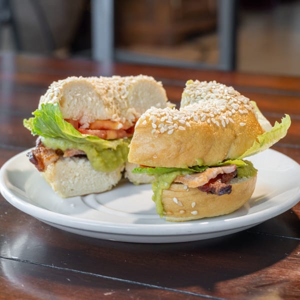 Two sesame-seed sandwiches with lettuce, tomato, avocado, and bacon served on a white plate atop a wooden table.