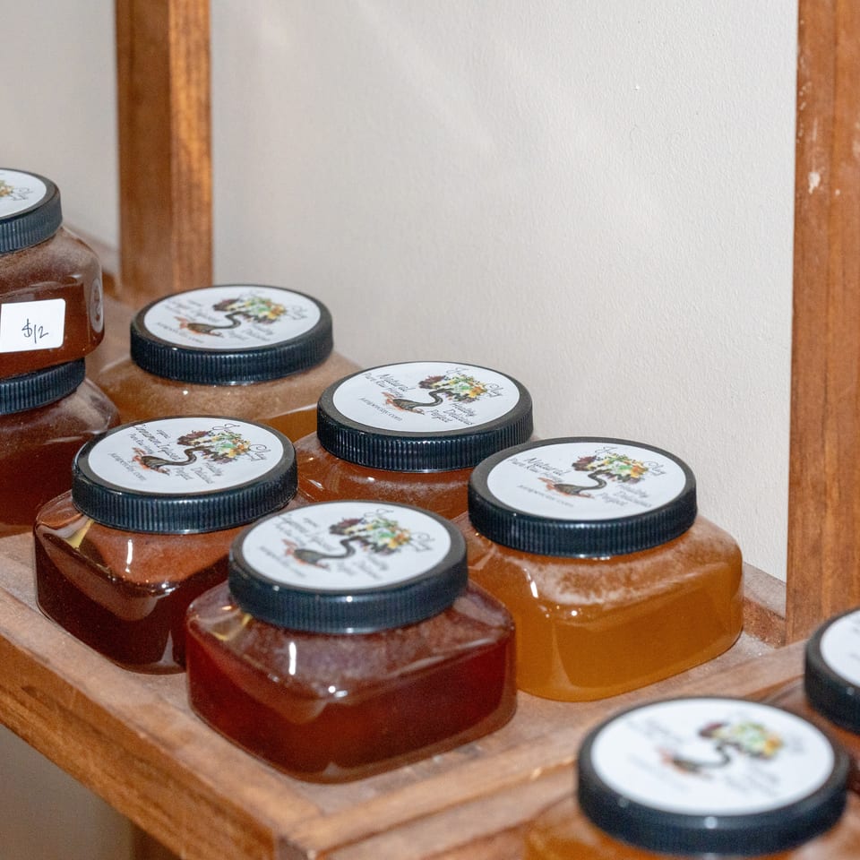 Rows of square glass jars filled with amber and red preserves sit on wooden shelves, featuring illustrated white labels and black lids.