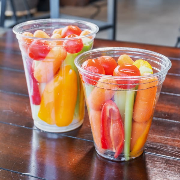 Two clear cups filled with colorful fresh fruit and vegetables sit on a wooden restaurant table.