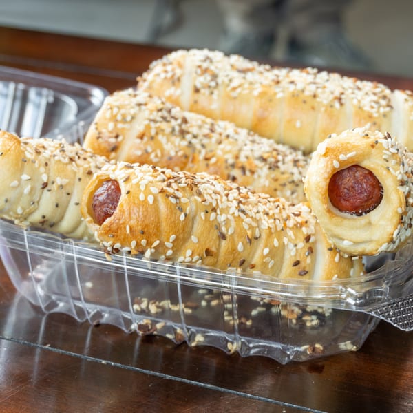Golden, sesame- and seed-covered sausage rolls arranged in a clear serving tray on a wooden table.