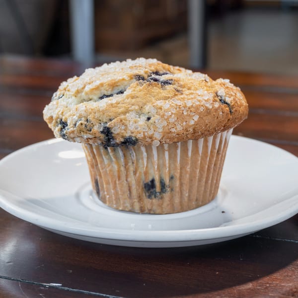 Blueberry muffin topped with coarse sugar, served on a white plate atop a wooden table.