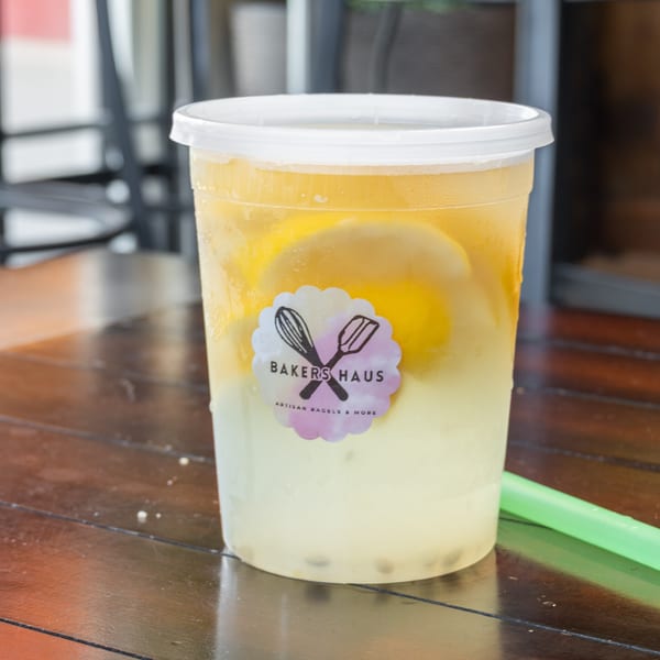 Clear cup of yellow citrus drink with lemon slices, Bakers Haus logo, and green straw on a wooden table.