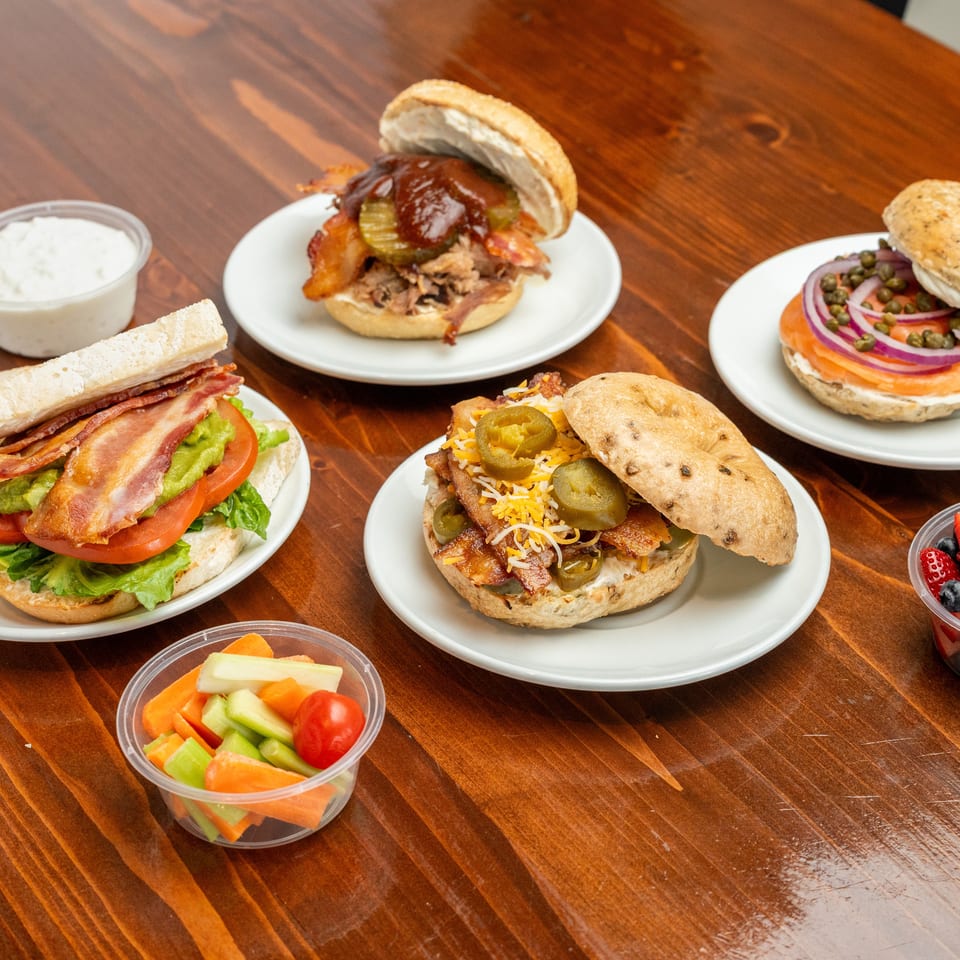 Assortment of loaded sandwiches, burgers, dipping sauce, fresh fruit, and vegetable sticks arranged on plates across a wooden table.