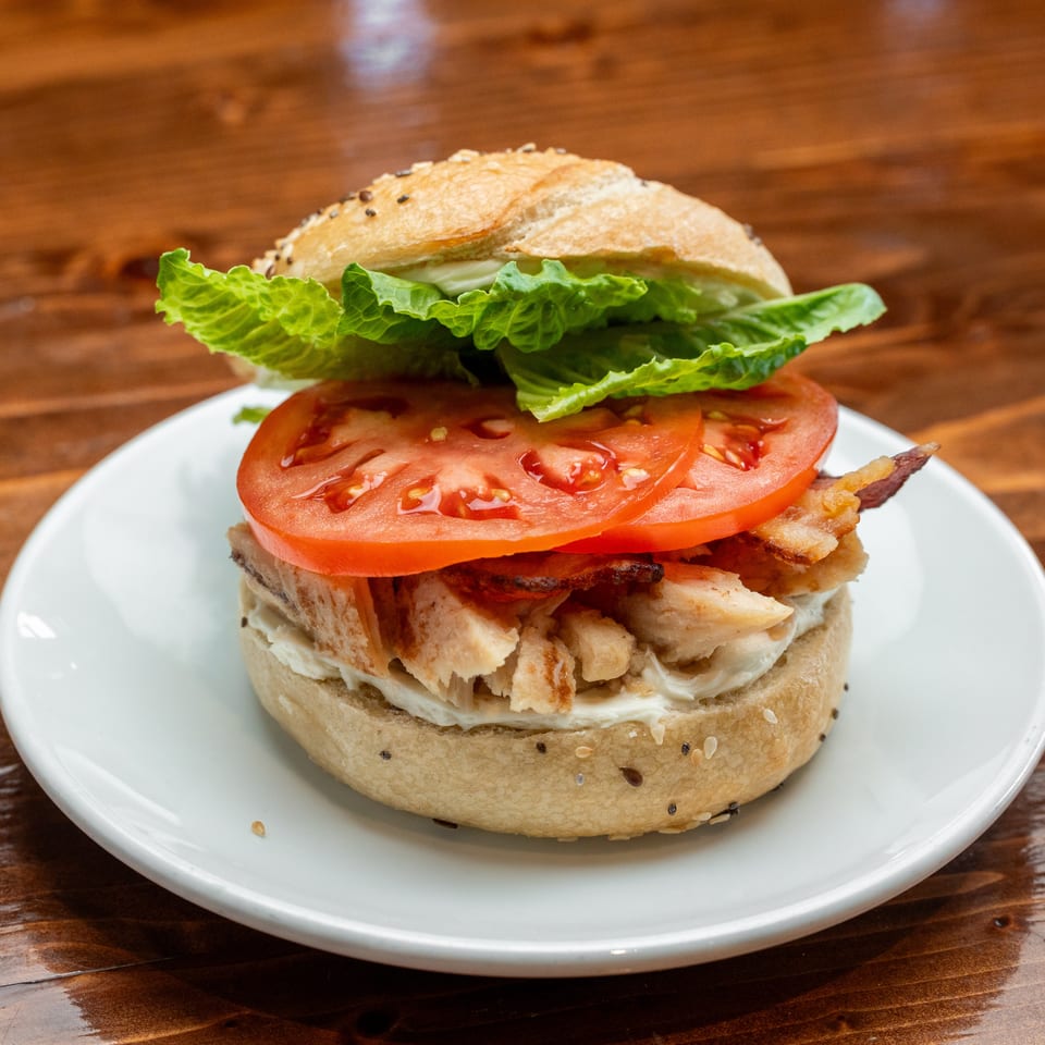 Chicken sandwich with lettuce, tomato, bacon, and creamy sauce on a seeded bun, served on a white plate.