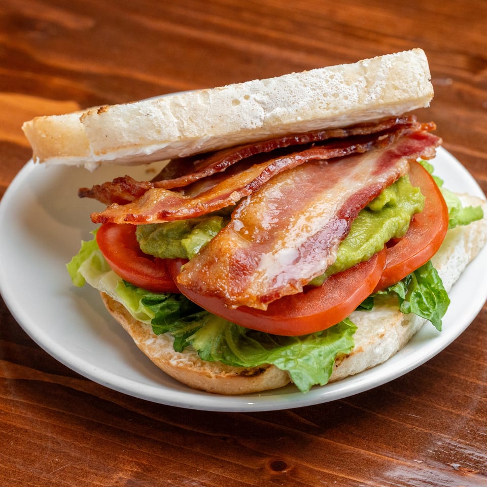 Bacon, lettuce, tomato, and avocado sandwich served on a white plate atop a wooden table.