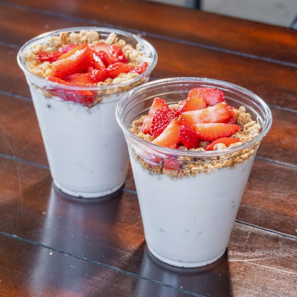Two clear cups of creamy yogurt topped with granola and sliced strawberries on a wooden table.