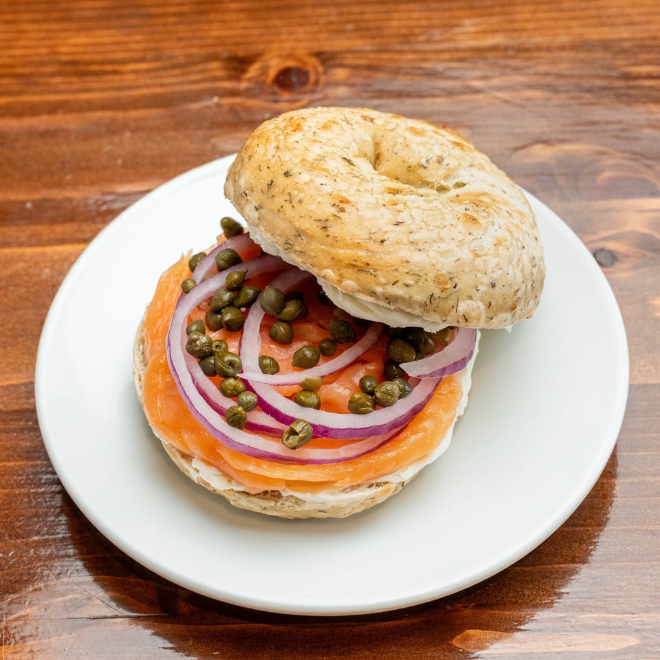 Bagel sandwich with smoked salmon, tomato, red onion, and capers on a white plate atop a wooden table.