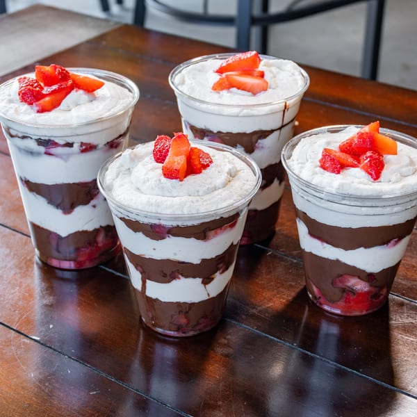 Five clear cups of layered chocolate, cream, and berries topped with sliced strawberries on a wooden table.