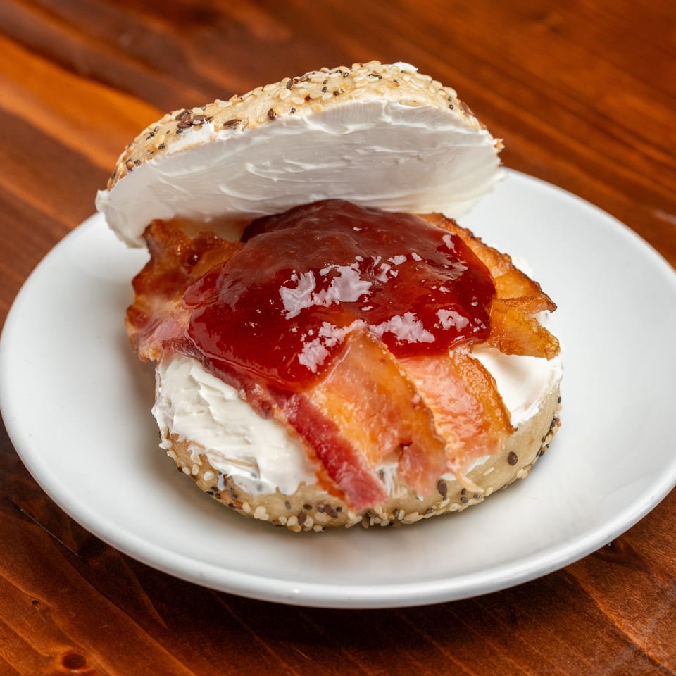 Sesame bagel sandwich with cream cheese, bacon, and glossy red jam on a white plate.