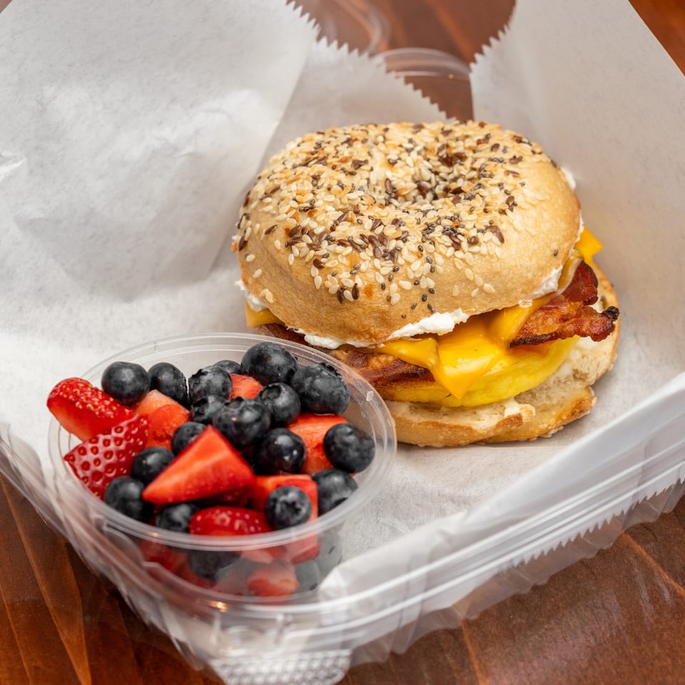 Everything bagel breakfast sandwich with bacon, egg, cheddar, and cream cheese beside a container of strawberries and blueberries.