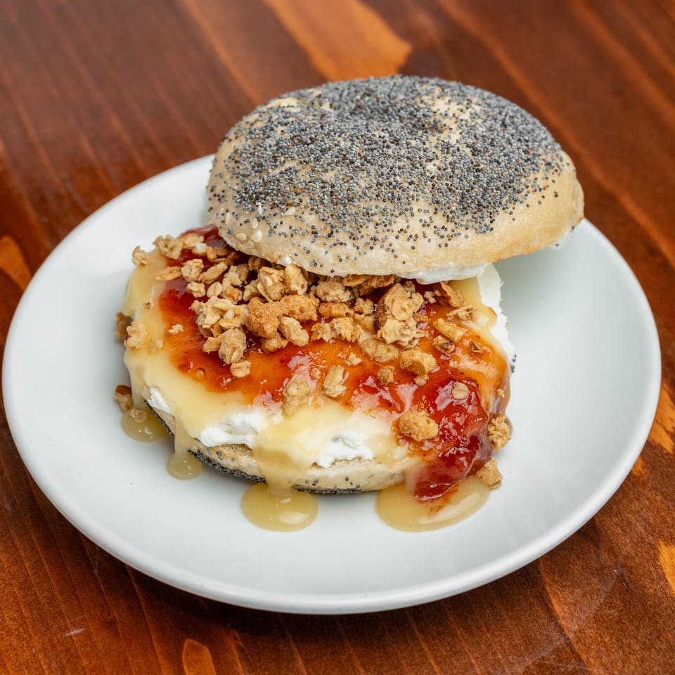 Bagel sandwich on a white plate, layered with cream cheese, red jam, crunchy granola, and honey on a wooden table.