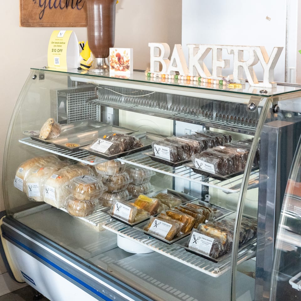 Bakery display case stocked with packaged brownies, breads, cakes, and pastries beneath a decorative bakery sign.