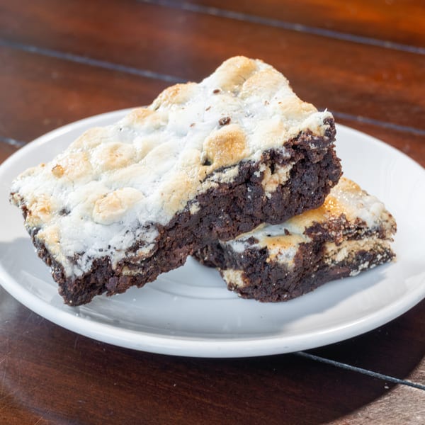 Stacked chocolate brownies topped with white frosting and chocolate chips on a white plate atop a wooden table.