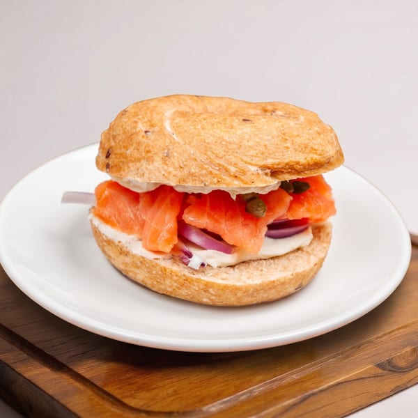 Bagel sandwich with smoked salmon, cream cheese, red onion, and capers on a white plate atop a wooden board.