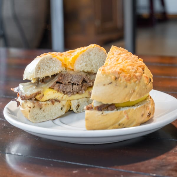 Two stacked meat and cheese sandwiches on toasted bread, served on a white plate atop a wooden restaurant table.